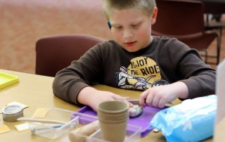 A child is working with clay, with materials such as picks and seeds spread out on the table in front of them.